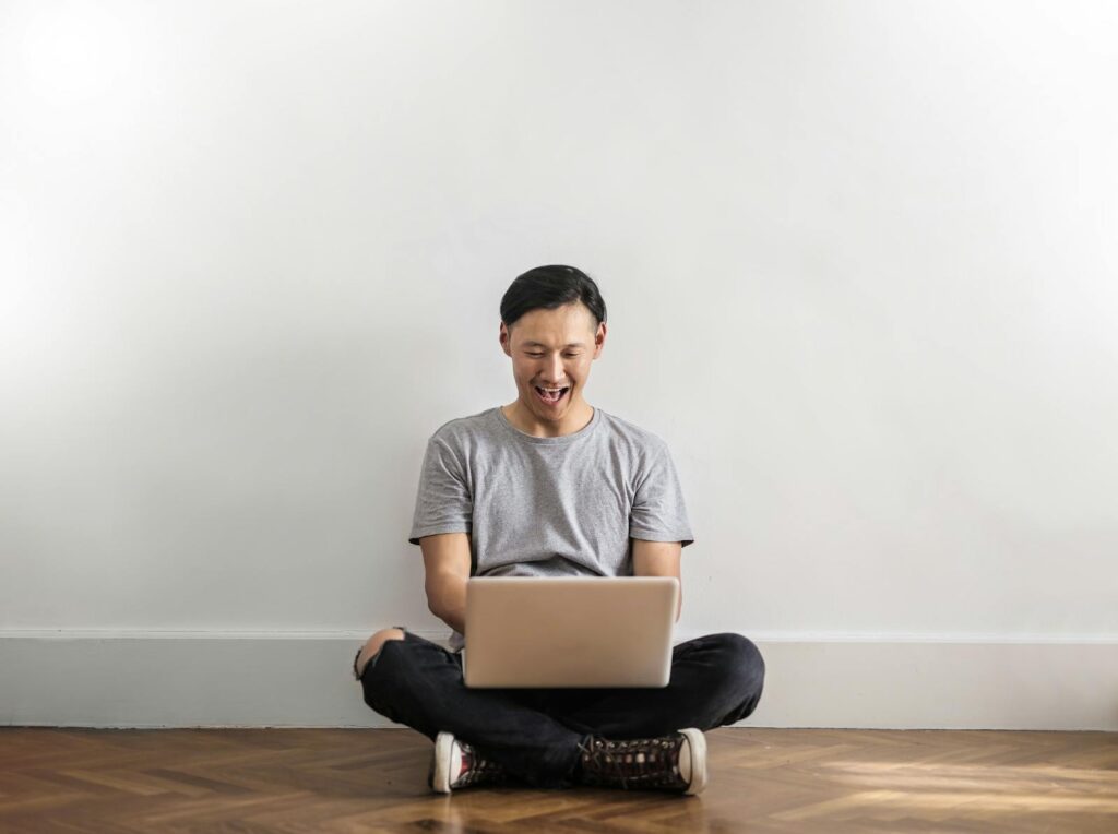 Photo of Laughing Man in Gray T-shirt and Black Jeans on Sitting on Wooden Floor While Using a Laptop