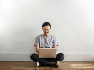 Photo of Laughing Man in Gray T-shirt and Black Jeans on Sitting on Wooden Floor While Using a Laptop